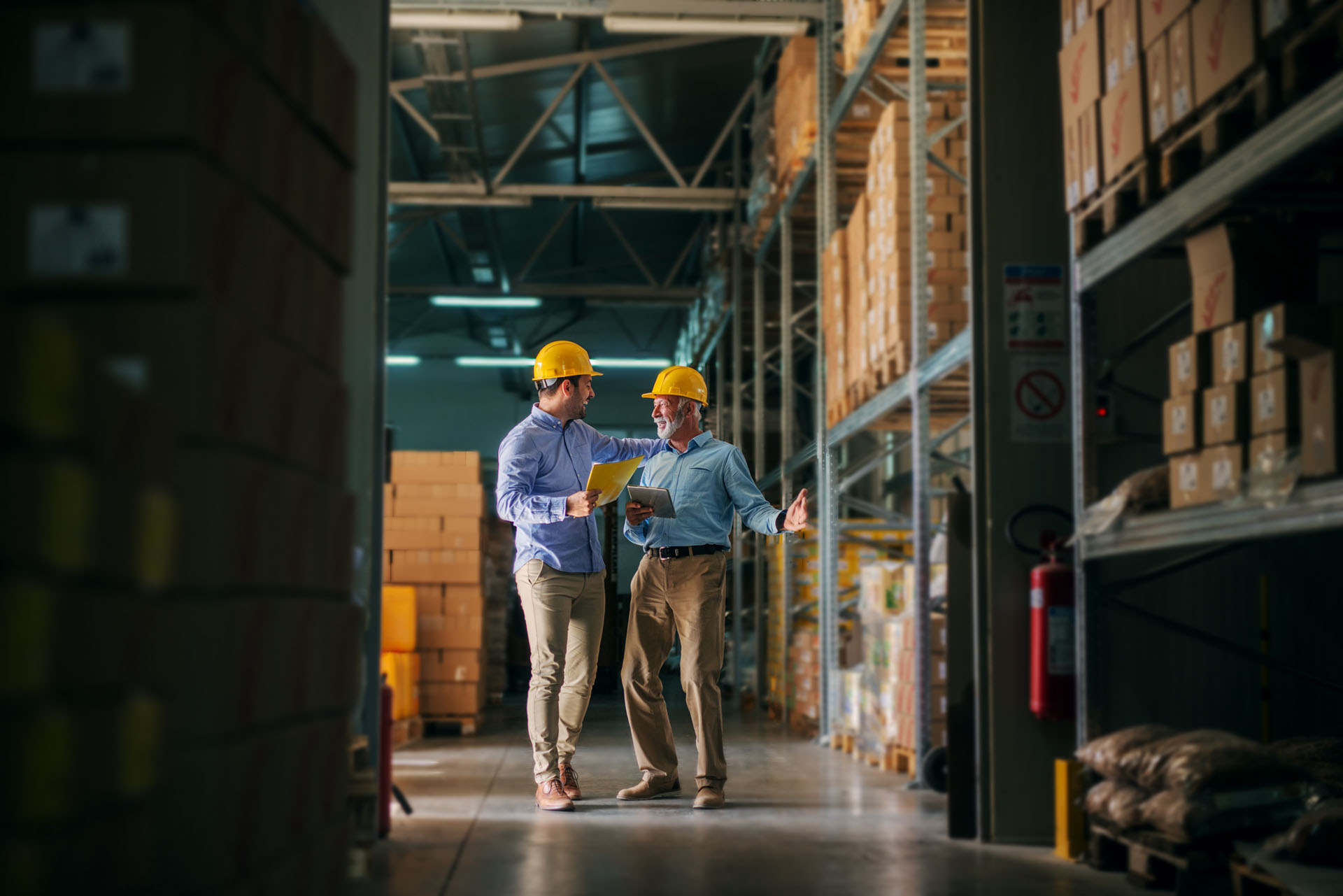 Picture of two business man standing in warehouse with helmets on their heads and celebrating great news about their business. Standing in big warehouse and looking happy and satisfied.